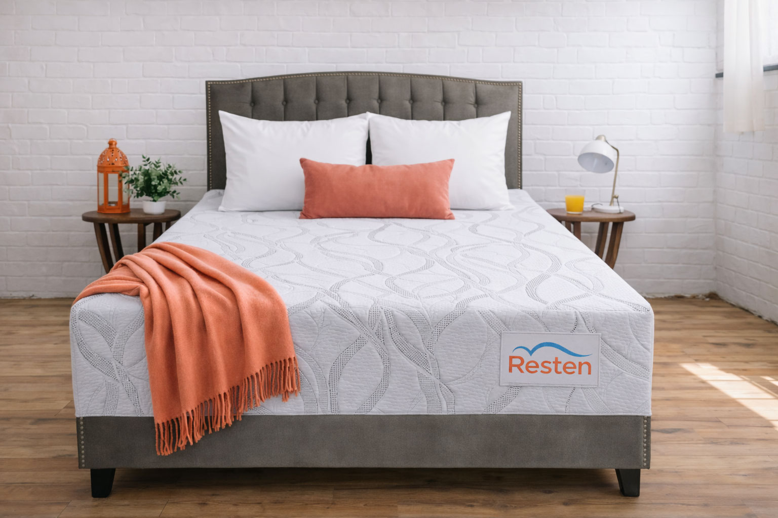 A Resten mattress with orange throw blanket and decorative pillow in a clean room with a white brick wall and wood flooring