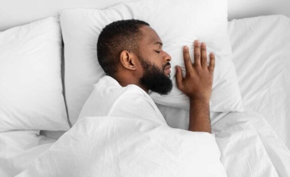 Top view of an all white bed and man sleeping on his side with his hand resting on the pillow beside his face.